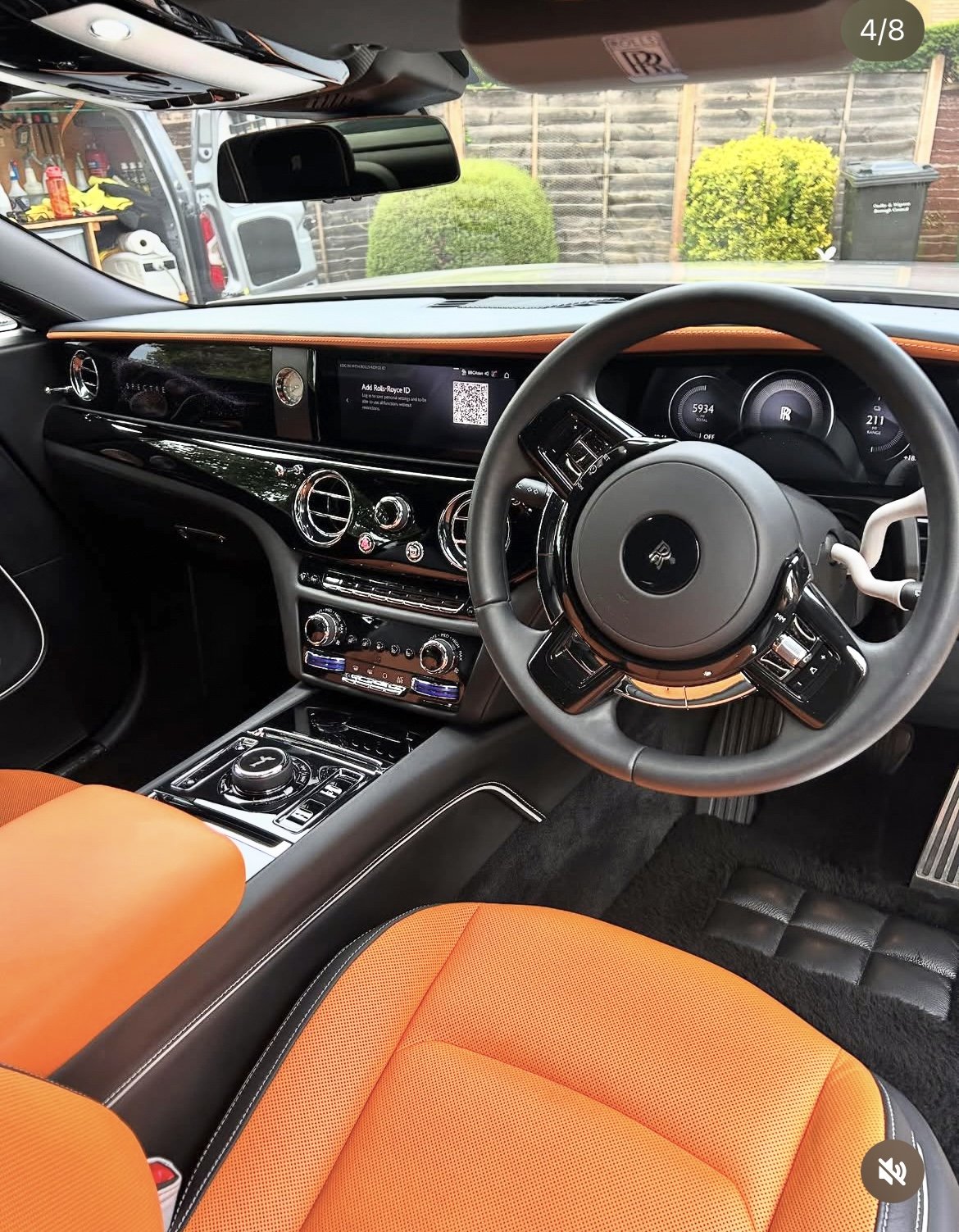 Interior of a luxury car, featuring a black dashboard with a digital display, a black steering wheel with the Rolls-Royce logo, orange seats, and a gear shift. Outside, a garden with green bushes and a compost bin are visible.
