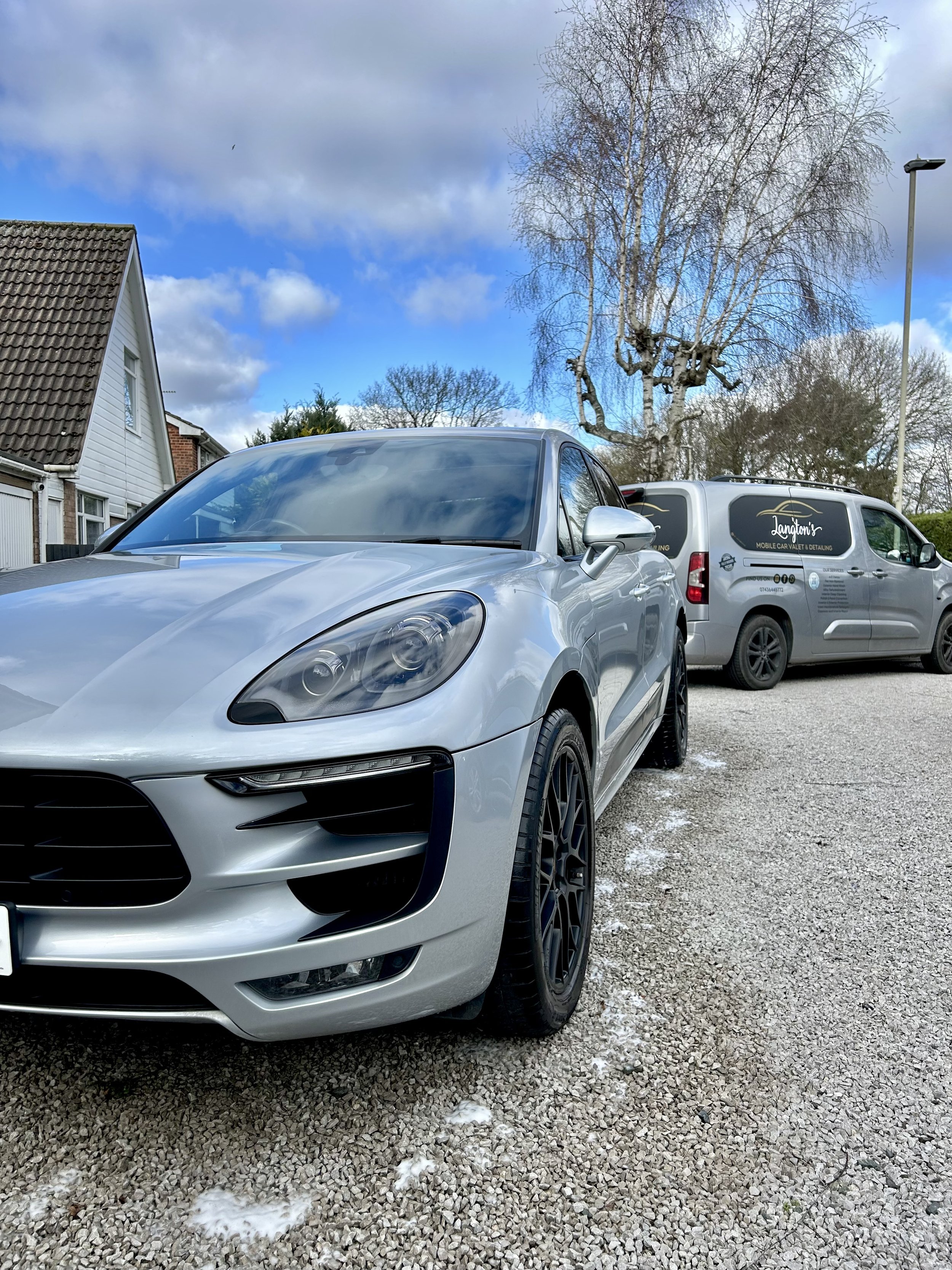 A silver sports car parked on a gravel driveway with a service van in the background and a partly cloudy sky overhead.
