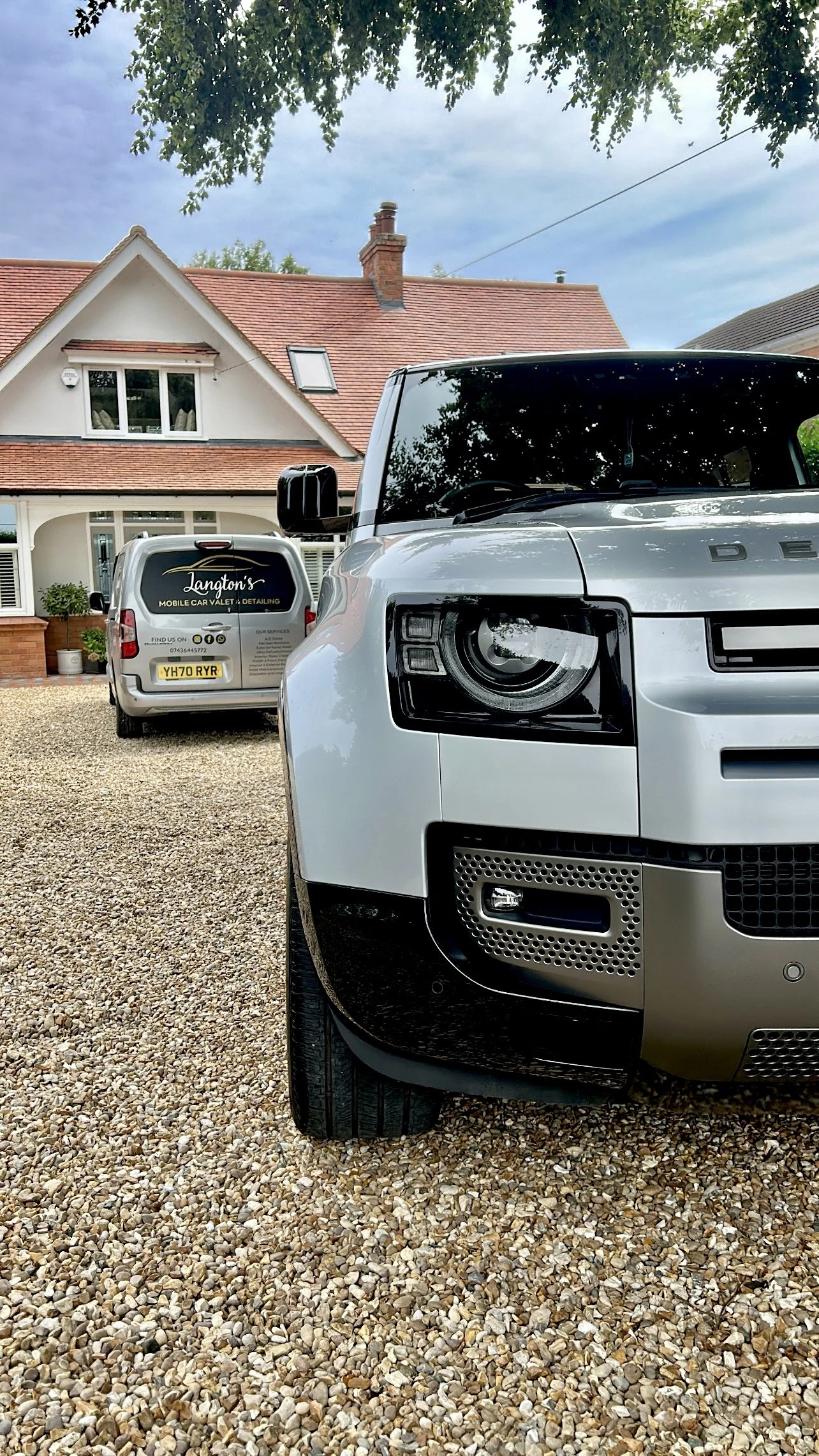 Close-up of a white Land Rover parked on a gravel driveway in front of a house with a red tiled roof and a second vehicle with branding for a mobile car valet service.