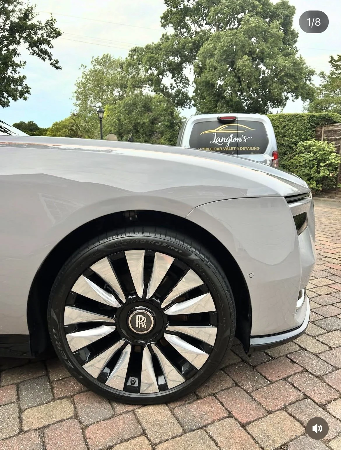 Close-up of a silver luxury car with a black and silver Rolls-Royce wheel rim, parked on a brick driveway with green trees in the background.