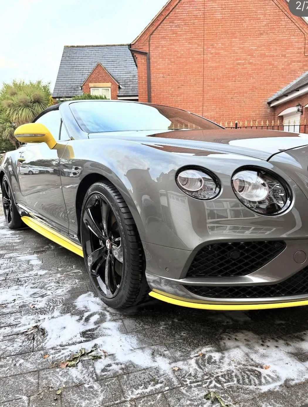 Gray sports car with yellow accents parked on wet driveway in front of red brick houses.
