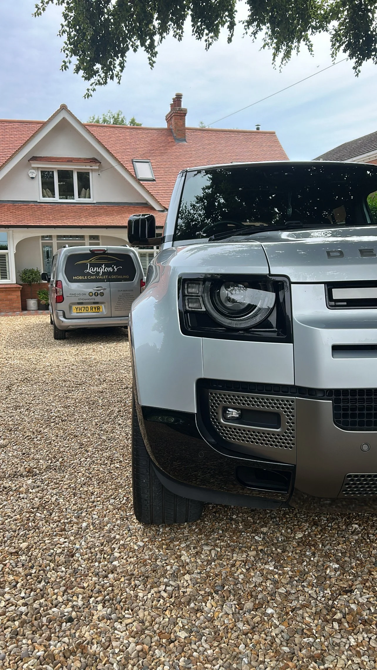 Close-up of the front of a silver Land Rover vehicle parked on a gravel driveway in front of a residential house with a red-tile roof. Another car with a valet company logo is parked in the background.