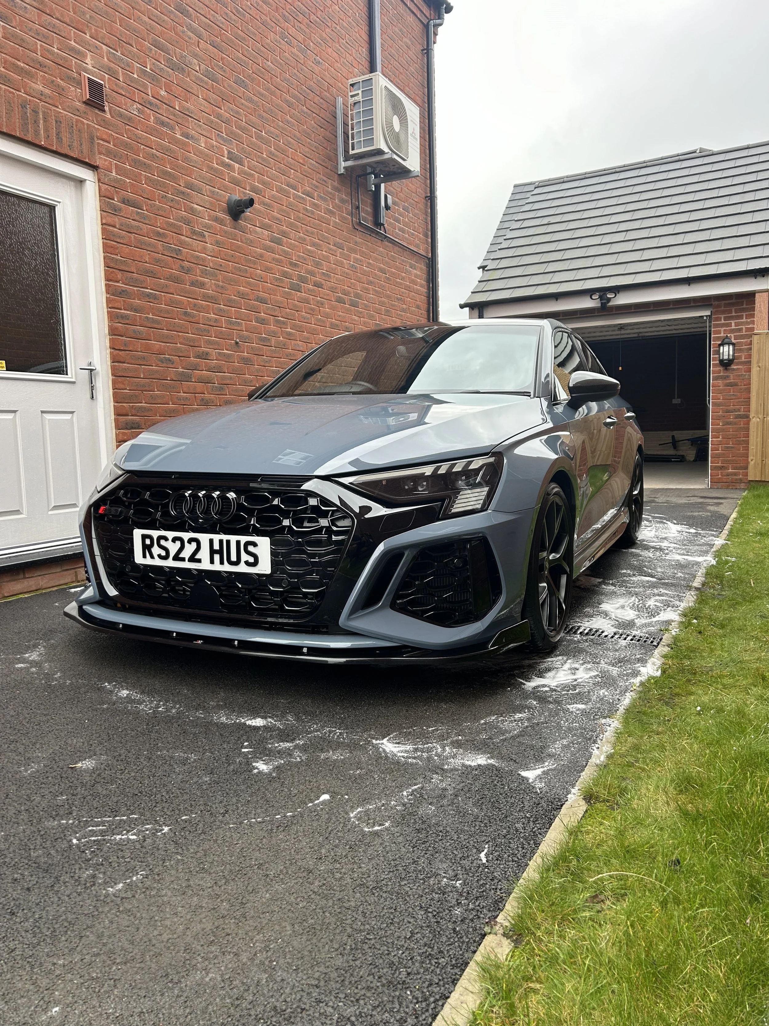 A gray Audi RS5 parked on a driveway with soap suds around it, next to a brick house with a garage and an air conditioning unit mounted on the wall.