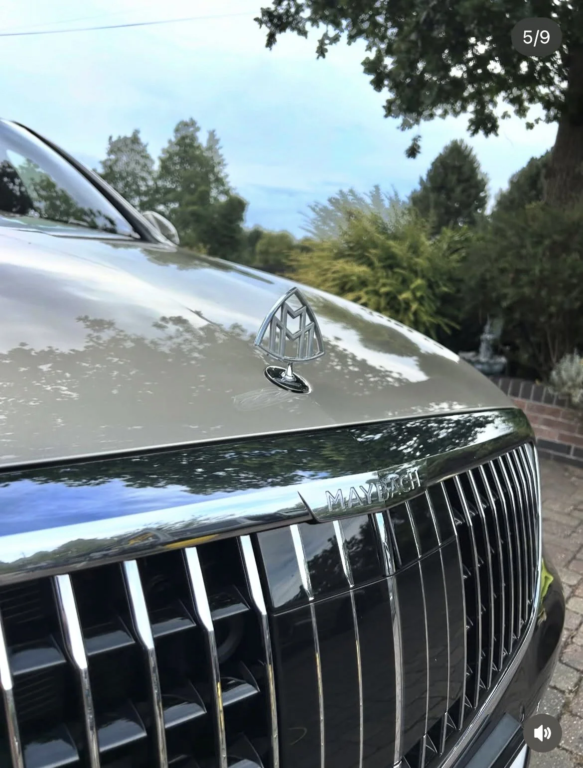 Close-up of a Maybach luxury car front, showing the chrome grille, Maybach emblem, and hood ornament, with trees and a blue sky in the background.