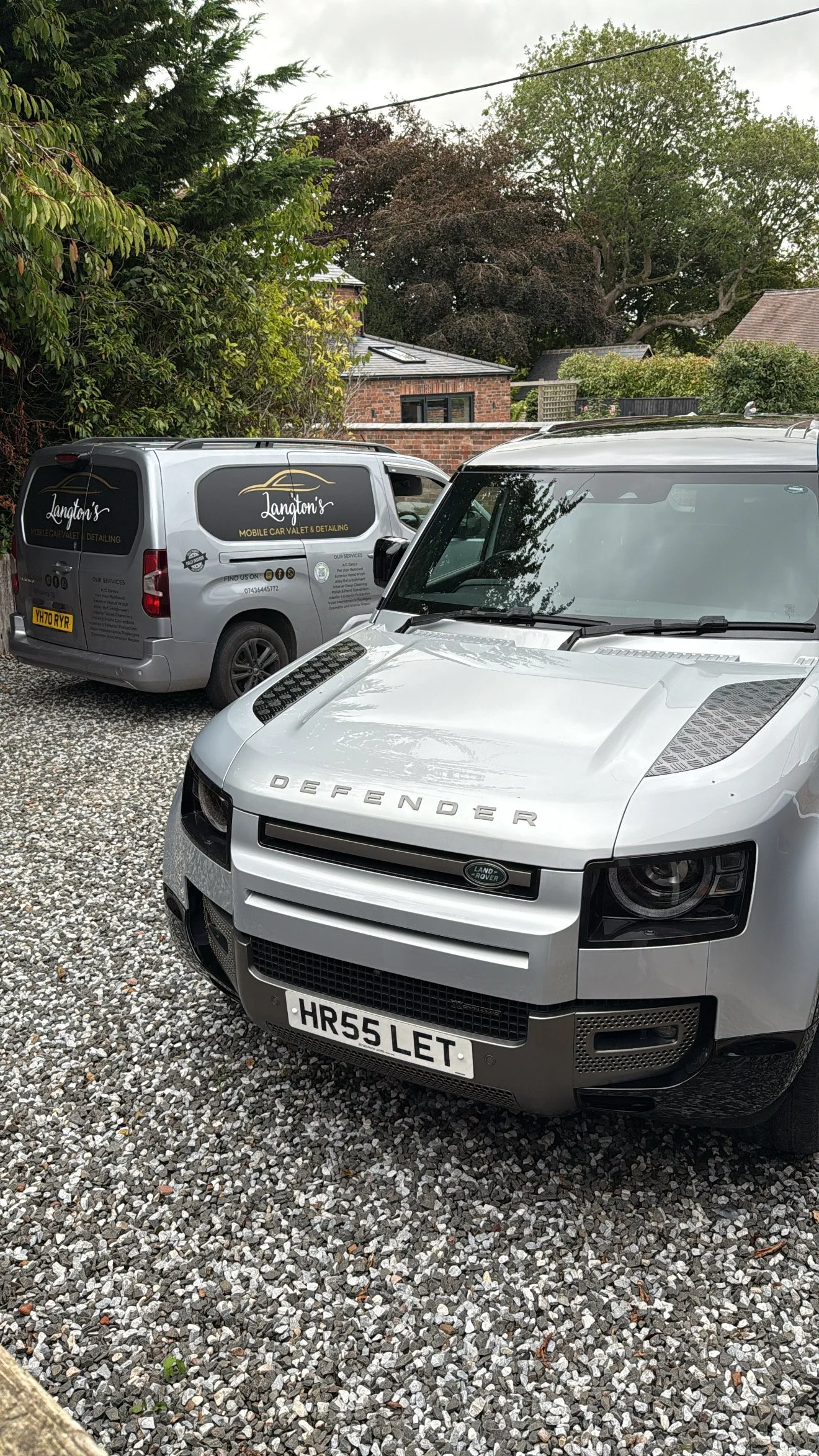 A silver Land Rover Defender parked on gravel, with a grey van labeled