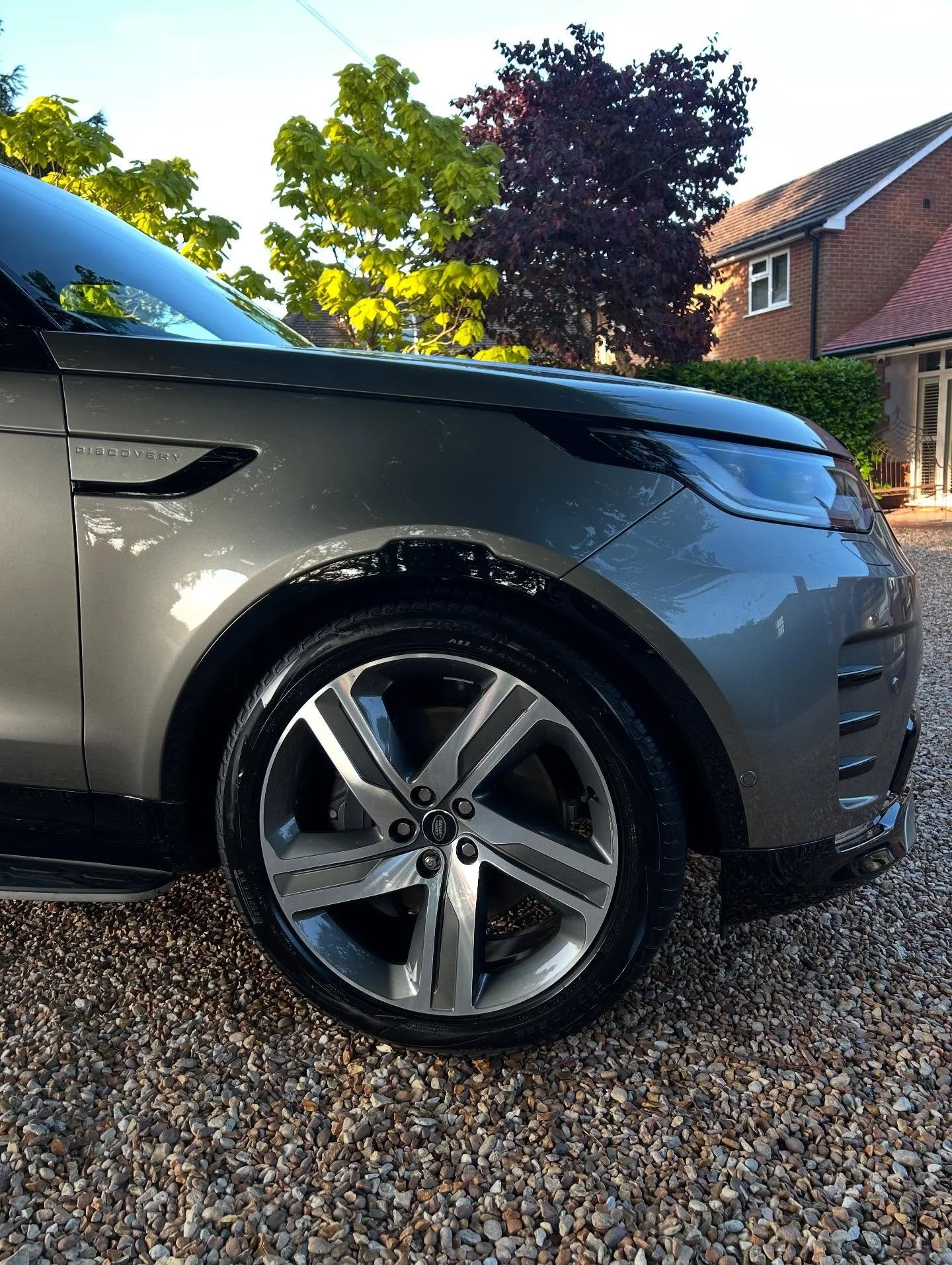 Close-up of the front left side of a gray Land Rover Discovery SUV parked on a gravel driveway with trees and a house in the background.