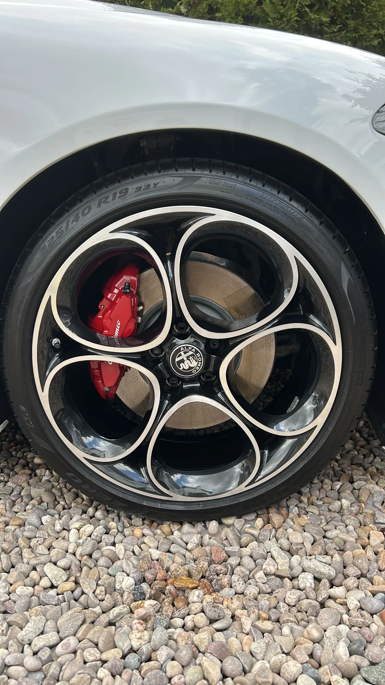 Close-up of a car wheel with a black and silver rim, a red brake caliper, and a black tire, parked on a gravel surface.