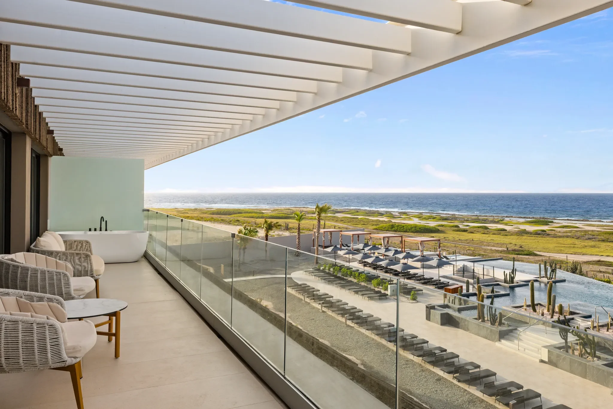 Balcón con sillones de mimbre, vista a la playa, área con tumbonas, parque con cactus, y el mar en el fondo.