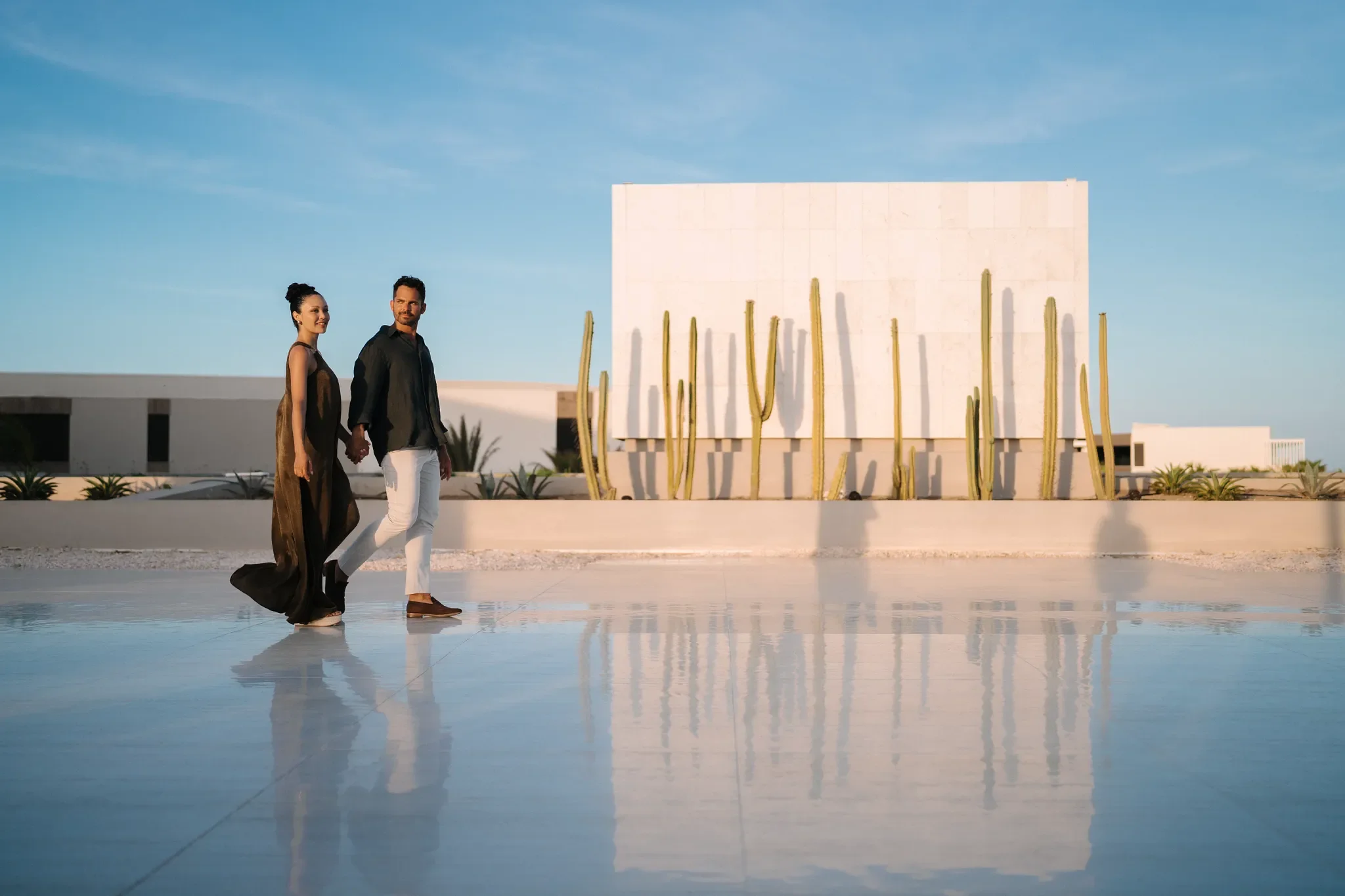 Una pareja caminando de la mano en un entorno moderno y minimalista al aire libre, con cactus y un edificio blanco al fondo, durante el día.