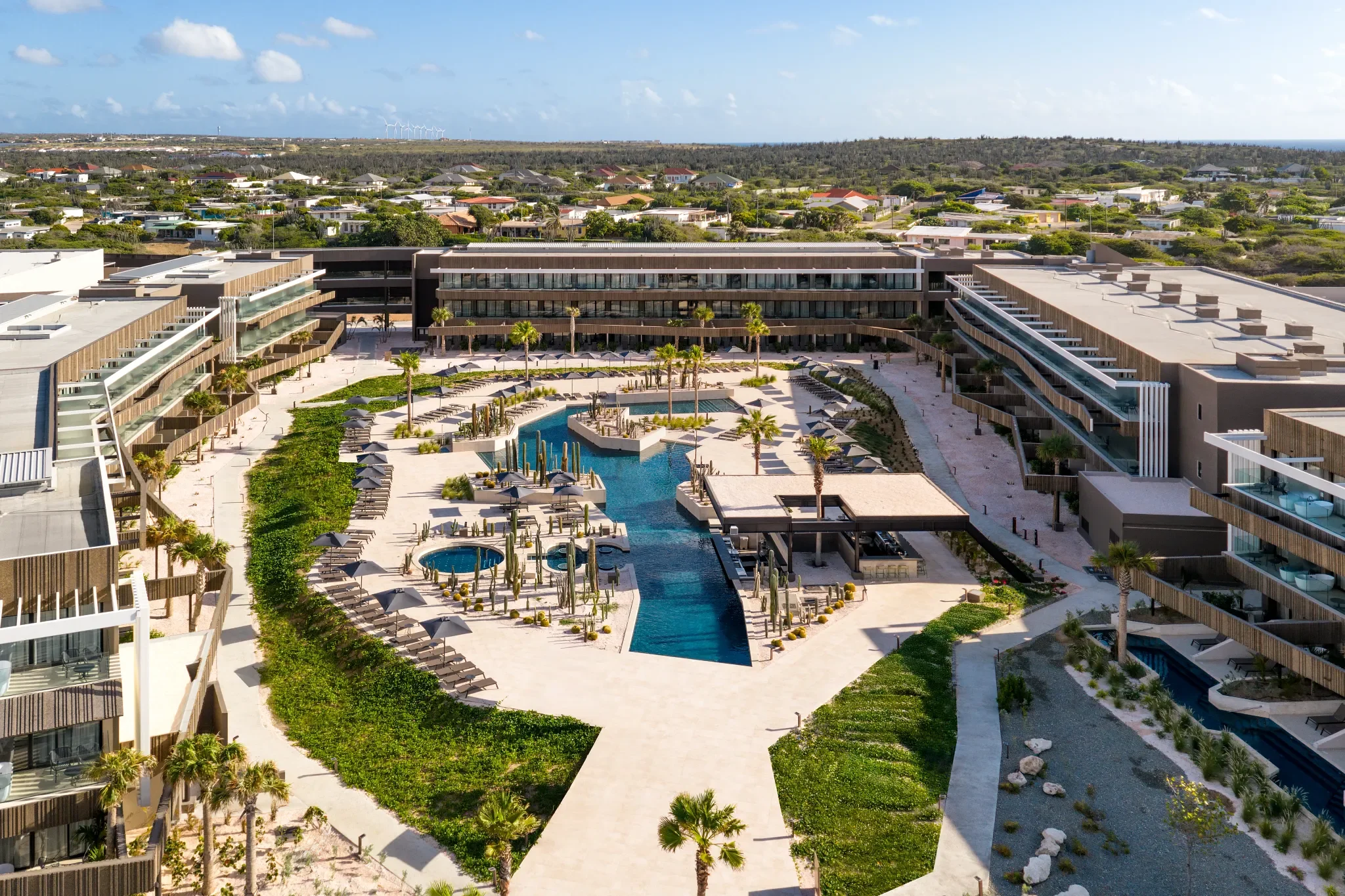 Vista aérea de un complejo hotelero con piscina central, áreas verdes y edificios modernos en un entorno soleado.
