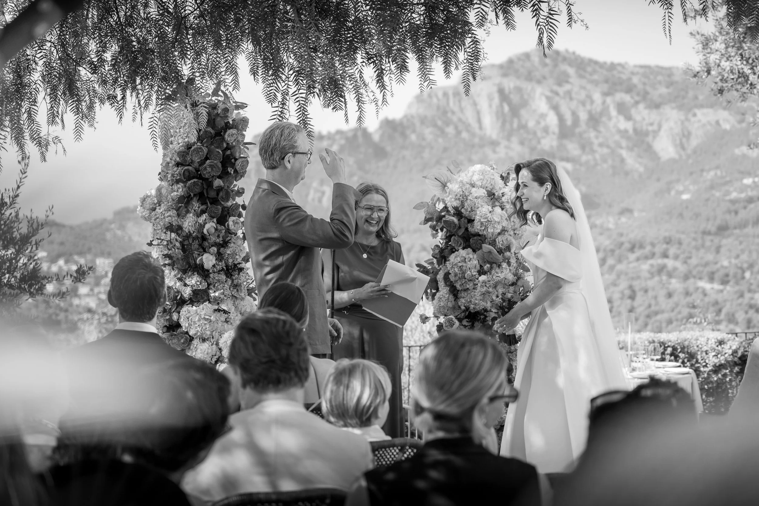 Una ceremonia matrimonial al aire libre con una pareja de novios sonriendo, rodeados de flores y asistentes, en un entorno natural con montañas de fondo.