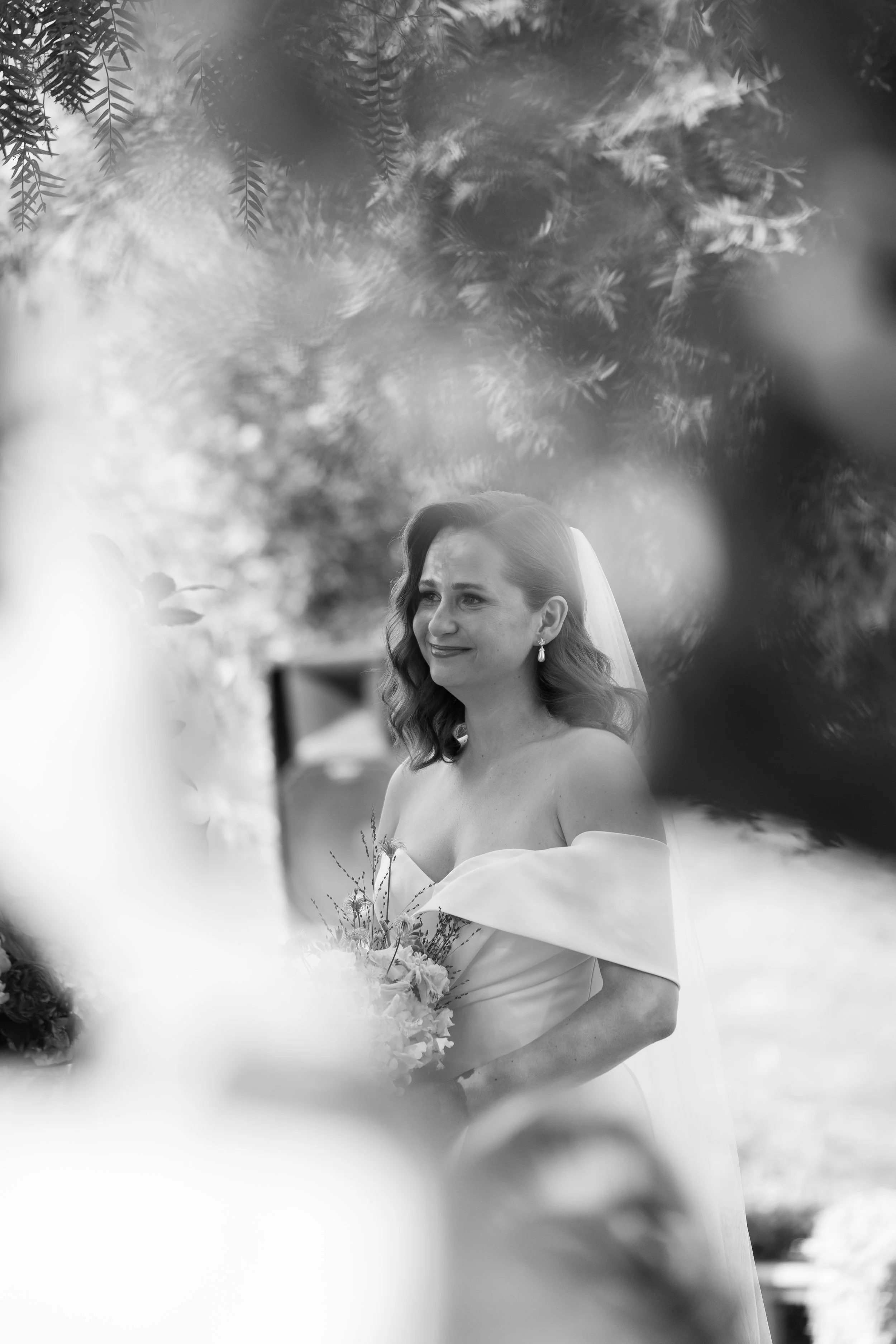 Mujer en vestido de novia sosteniendo un ramo de flores en un entorno al aire libre, con árboles y vegetación a su alrededor, en una fotografía en blanco y negro.