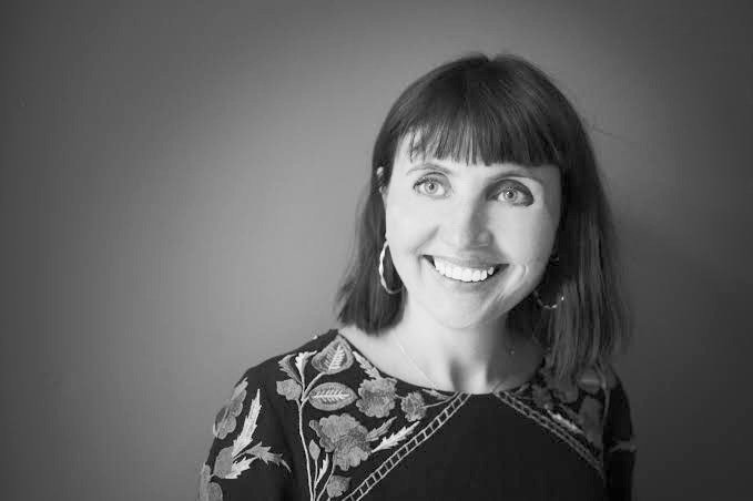 Black and white photo of a smiling woman with shoulder-length dark hair and bangs, wearing hoop earrings and a floral patterned top, looking slightly to the right.