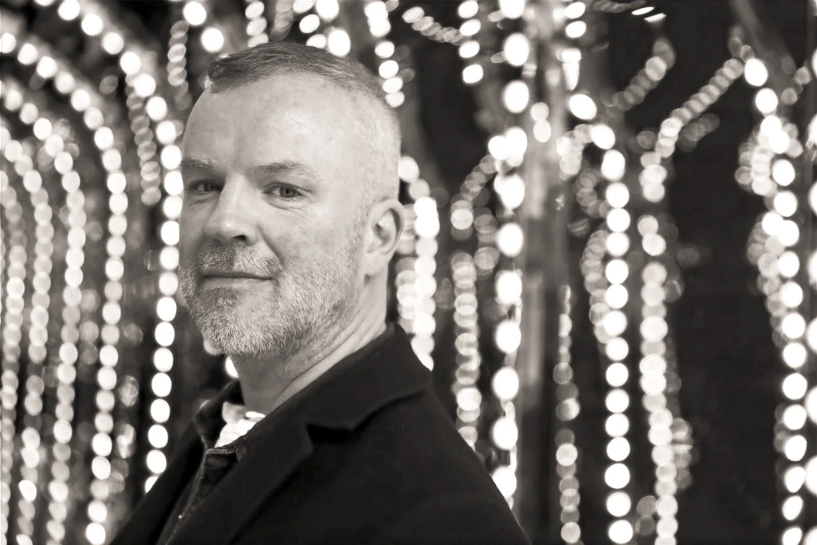 A man with short hair and beard dressed in a tuxedo, posing in front of a background of hanging lights or bokeh.