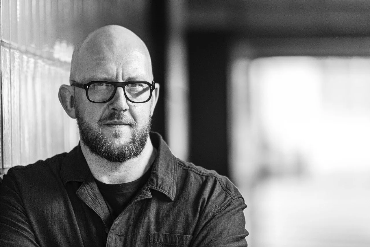 Black and white photo of a bald man with a beard and glasses, leaning against a wall with his arms crossed, looking at the camera.