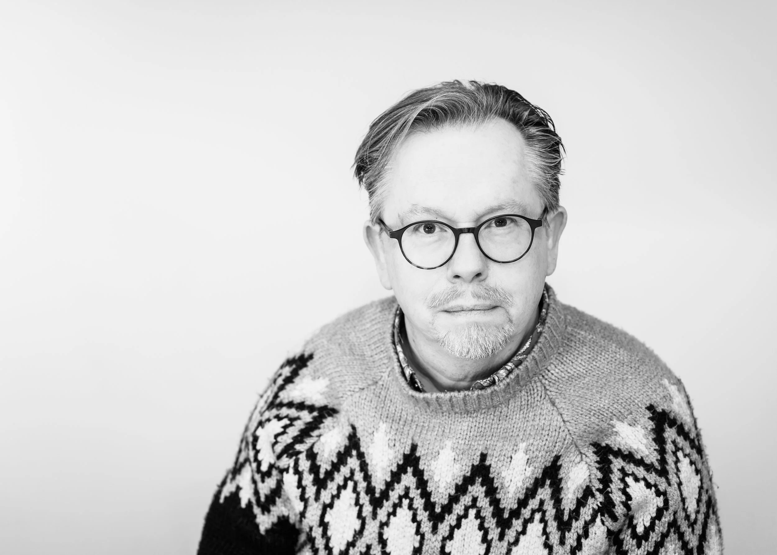 Black and white portrait of a man wearing glasses and a patterned sweater, looking at the camera with a neutral expression.