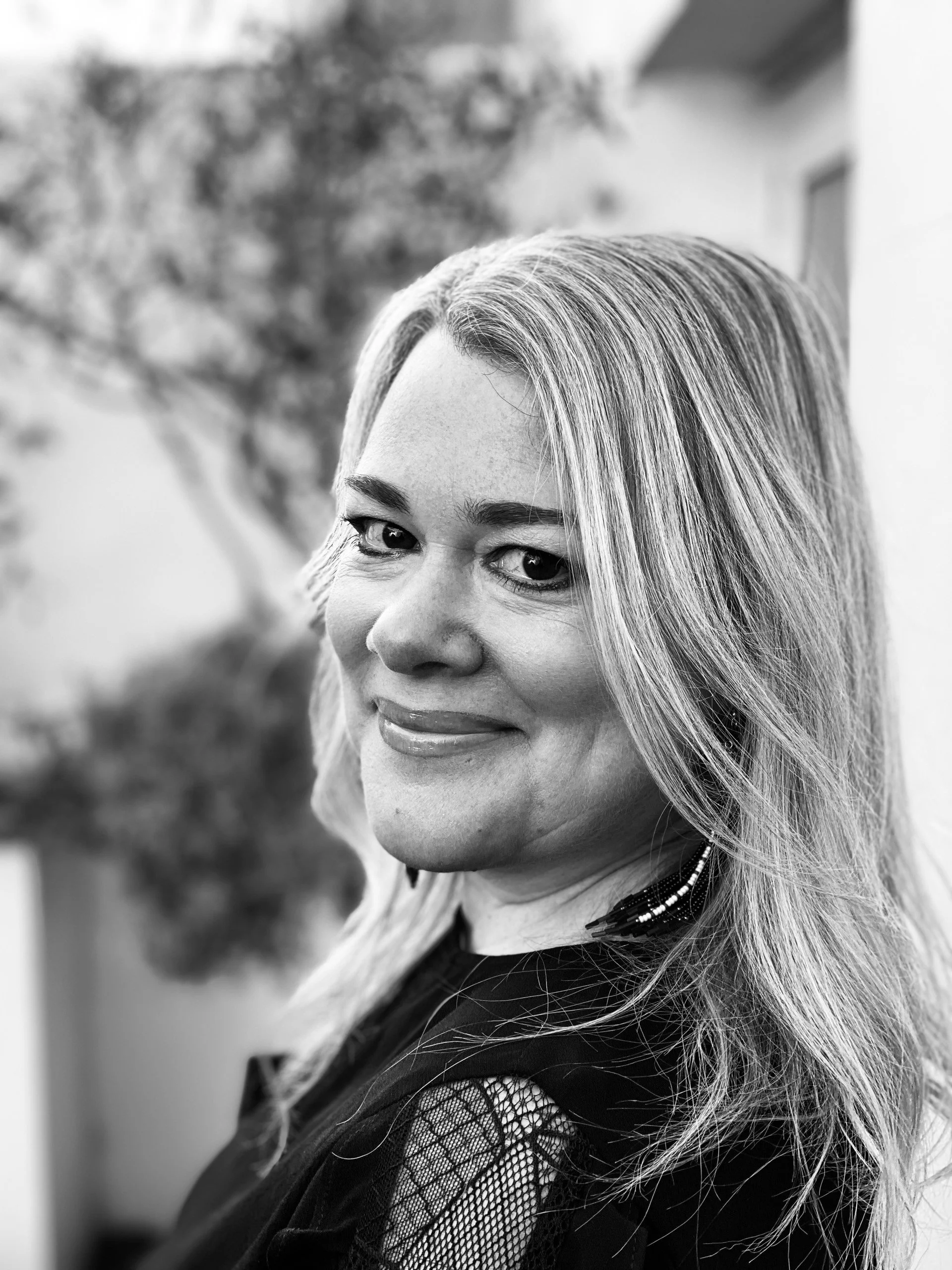 Black and white photo of a smiling woman with shoulder-length wavy hair, wearing earrings and a black top with lace details.