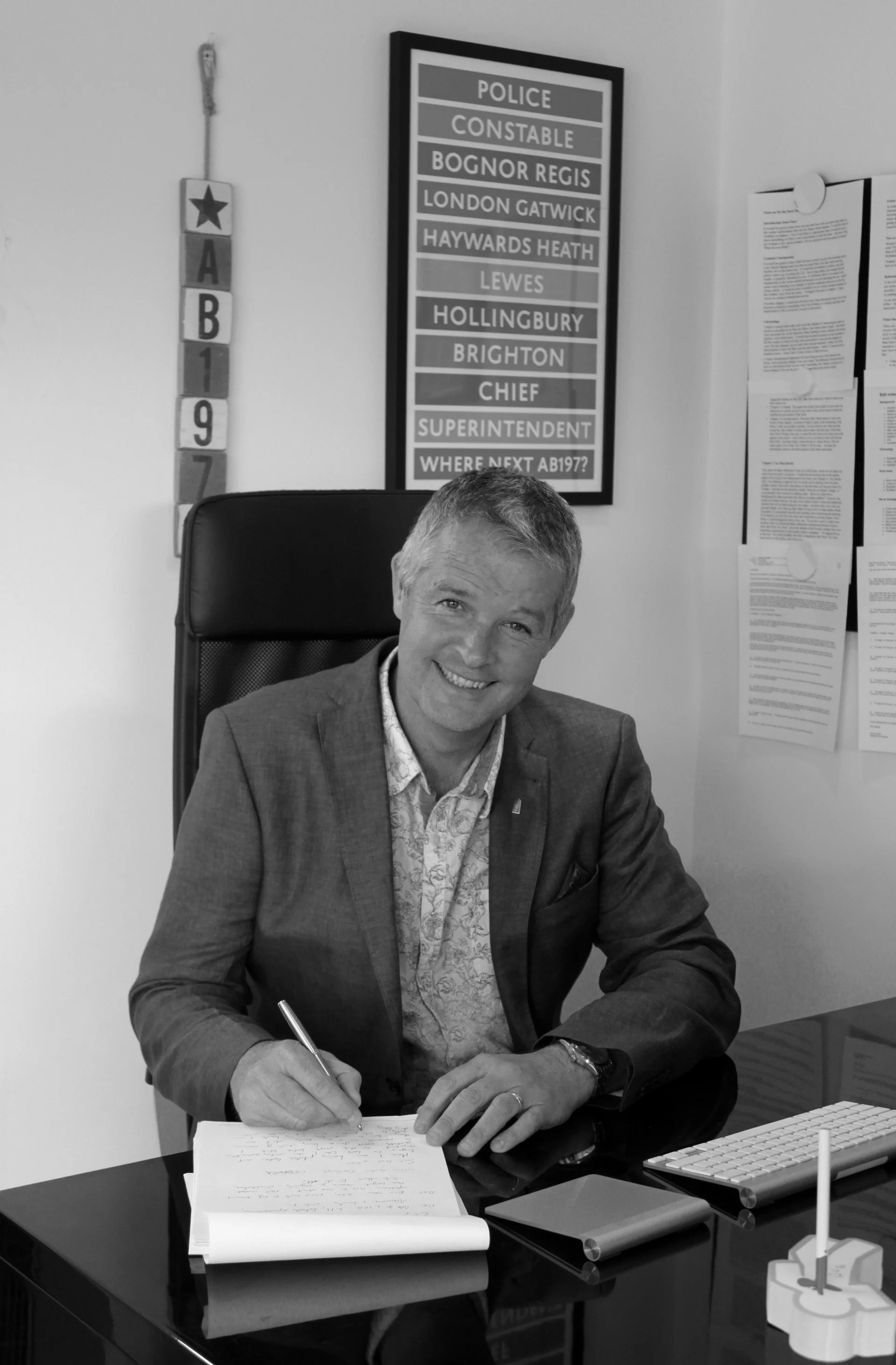 A man in a suit sitting at a desk, smiling, writing on a notepad with a pen. Behind him on the wall are a police badge with a star at the top, and a framed display with police station names and a question about the next police superintendent.