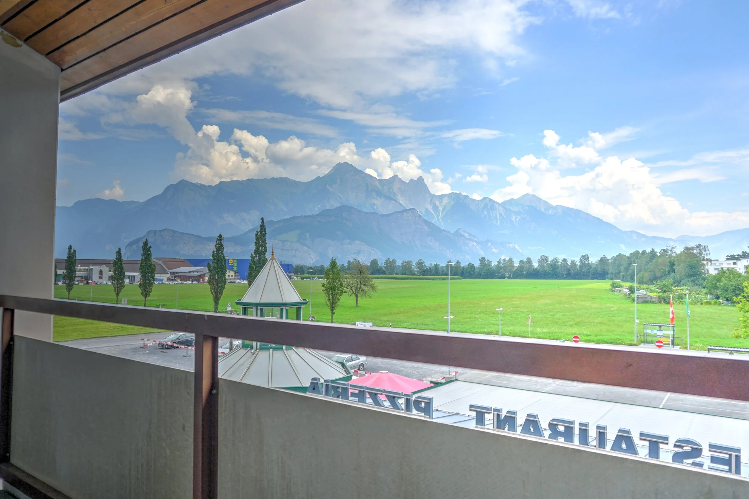 Blick aus einem Fenster auf eine grüne Wiese mit Bäumen, im Hintergrund Berge und Himmel mit Wolken.