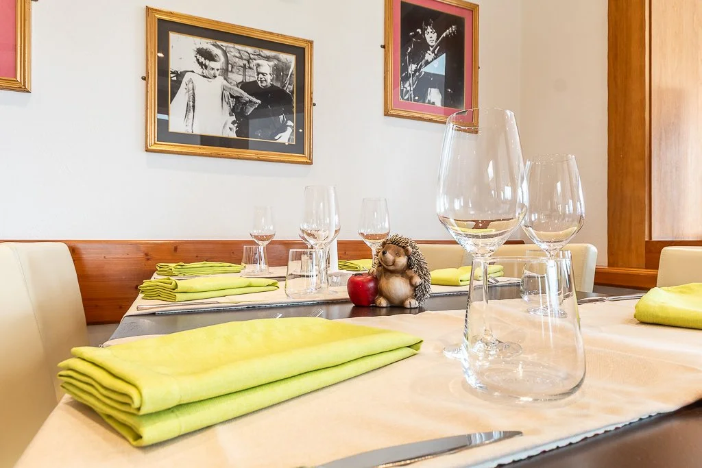 Ein festlich gedeckter Esstisch mit gelben Geschirrtüchern, mehreren Weingläsern, Wasserhähnen und einer kleinen niedlichen Figur eines Igels, der neben einem roten Apfel sitzt, in einem Restaurant mit Wandbildern.