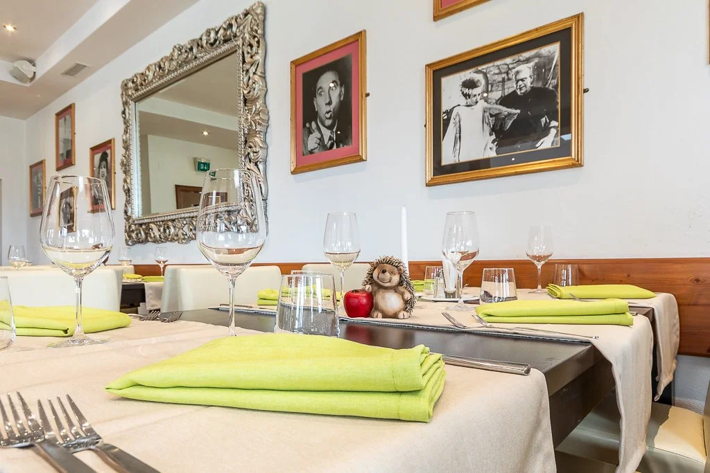 Ein Esstisch in einem Restaurant, gedeckt mit weißen Tischtüchern, grünen Servietten, Weingläsern, Wasserbechern, Geschirr, Silberbesteck und einer Dekoration aus einer kleinen Figur, einer roten Apfel und einer Kerze. Die Wände sind mit mehreren sch