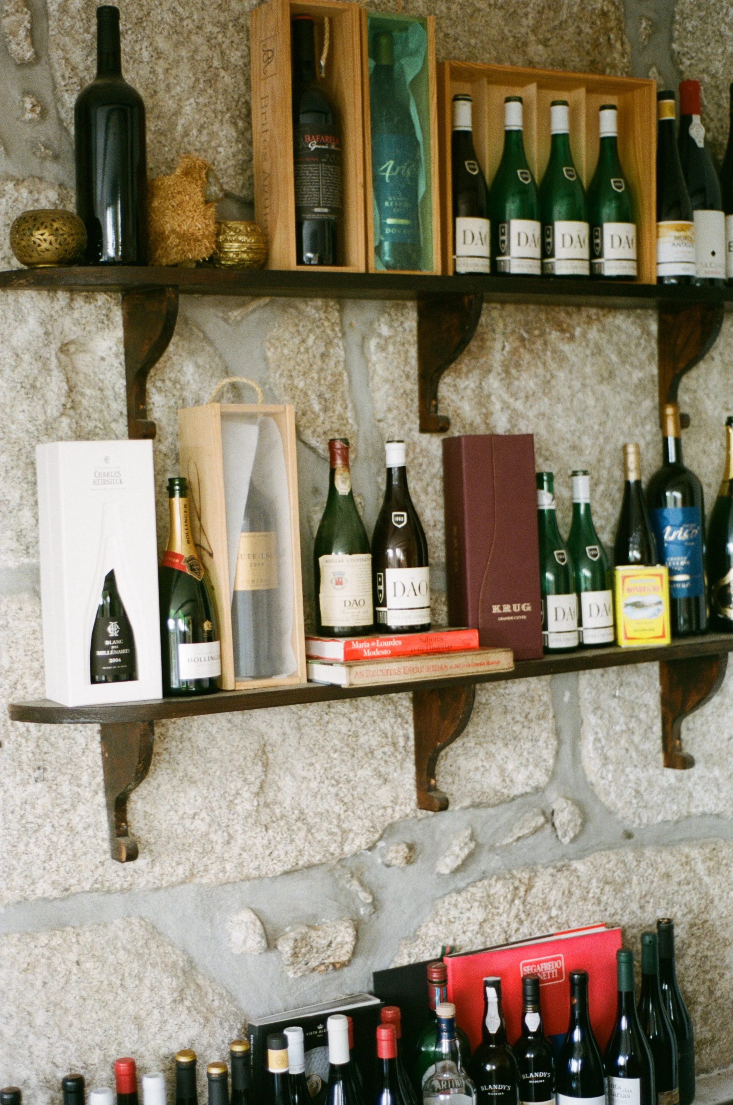 Vários tipos de garrafas de vinho e livros organizados em prateleiras de madeira e parede de pedra.
