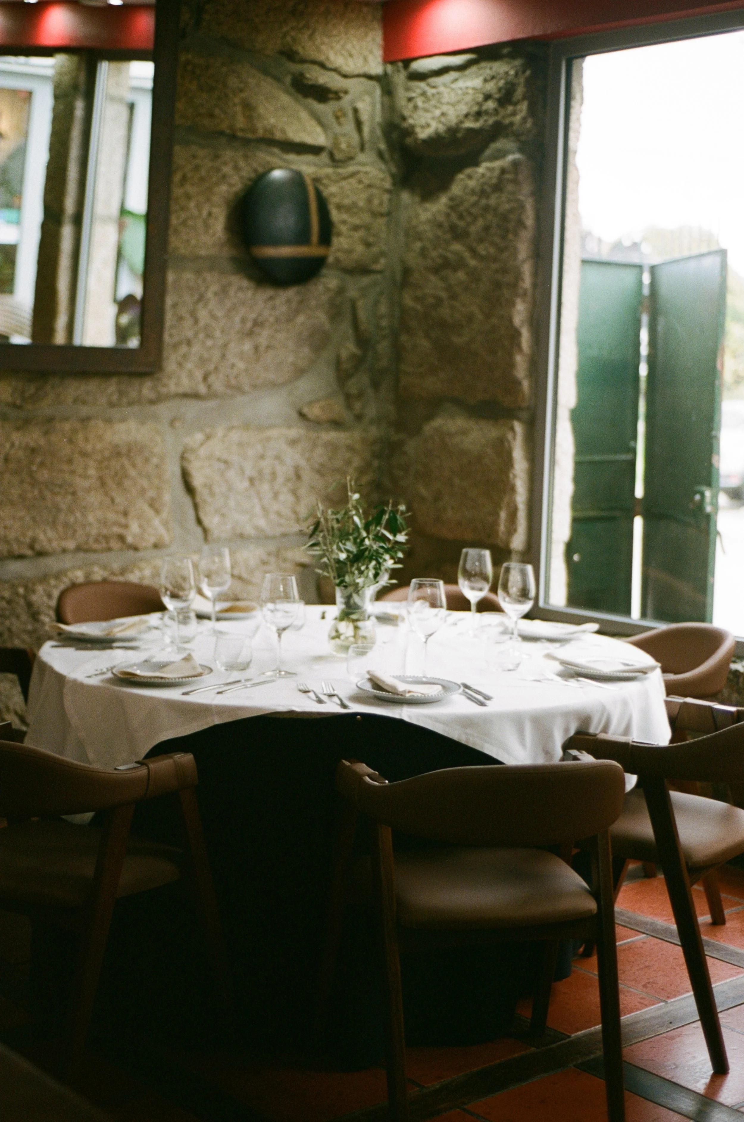 Mesa de jantar elegantemente arrumada com toalha branca, pratos, talheres, taças de vidro e um arranjo de flores no centro, em um ambiente com parede de pedra e janela aberta que deixa a luz natural entrar.