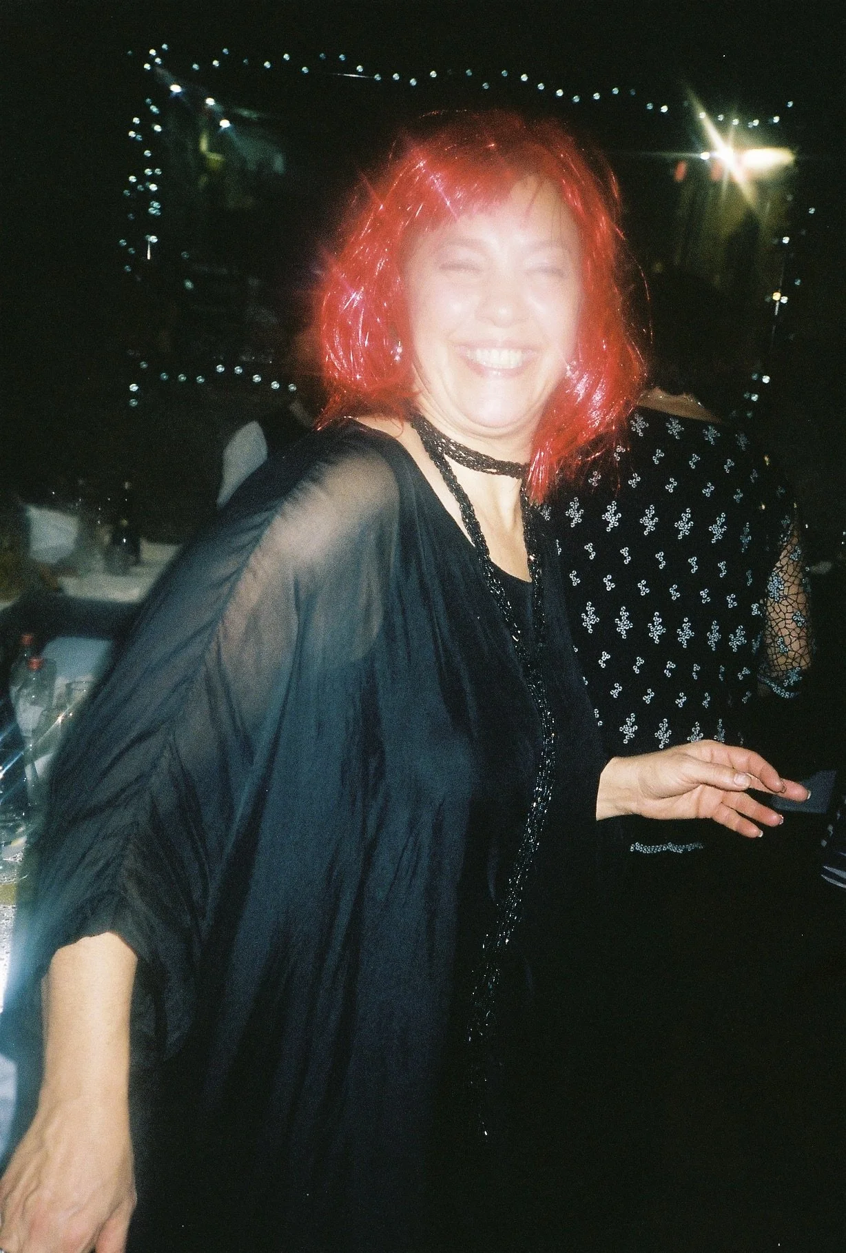 Mulher sorrindo com cabelo vermelho em ambiente de festa ou evento, usando roupa preta e acessório de colar
