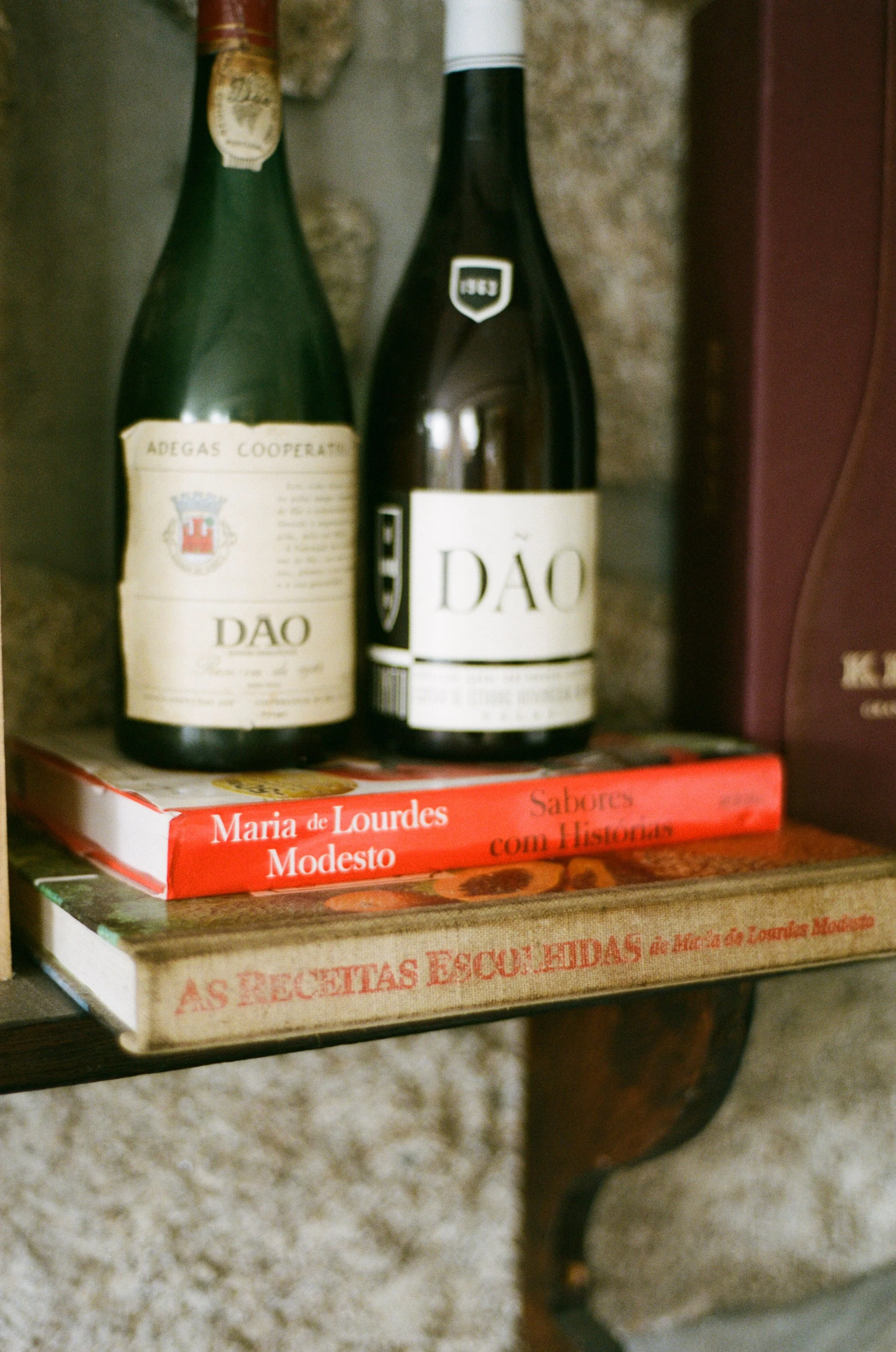 Duas garrafas de vinho em uma estante, ao lado de dois livros empilhados, um com capa vermelha chamado "Sabores com Histórias" de Maria de Lourdes Modesto, e outro com capa bege chamado "As Receitas Esquecidas" de Maria de Lourdes Modesto.