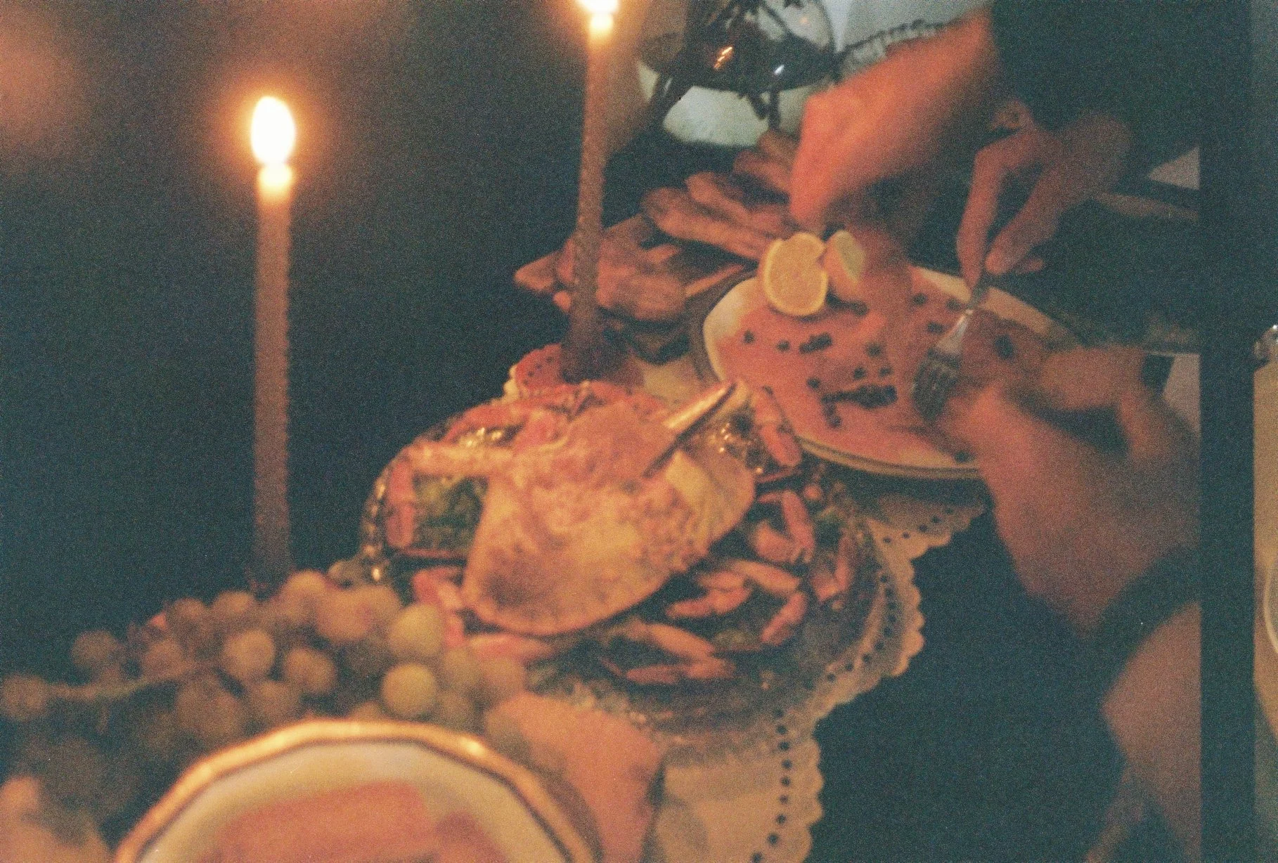 Refeição de aniversário com bolo decorado, frutas, uvas, e uma vela acesa ao lado, em uma mesa com uma toalha branca. Pessoas celebrando, com uma mão segurando um garfo.