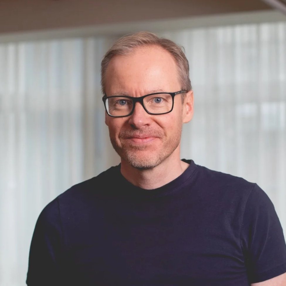 A middle-aged man with glasses and light-colored hair wearing a black T-shirt, smiling at the camera in front of a window with white curtains.