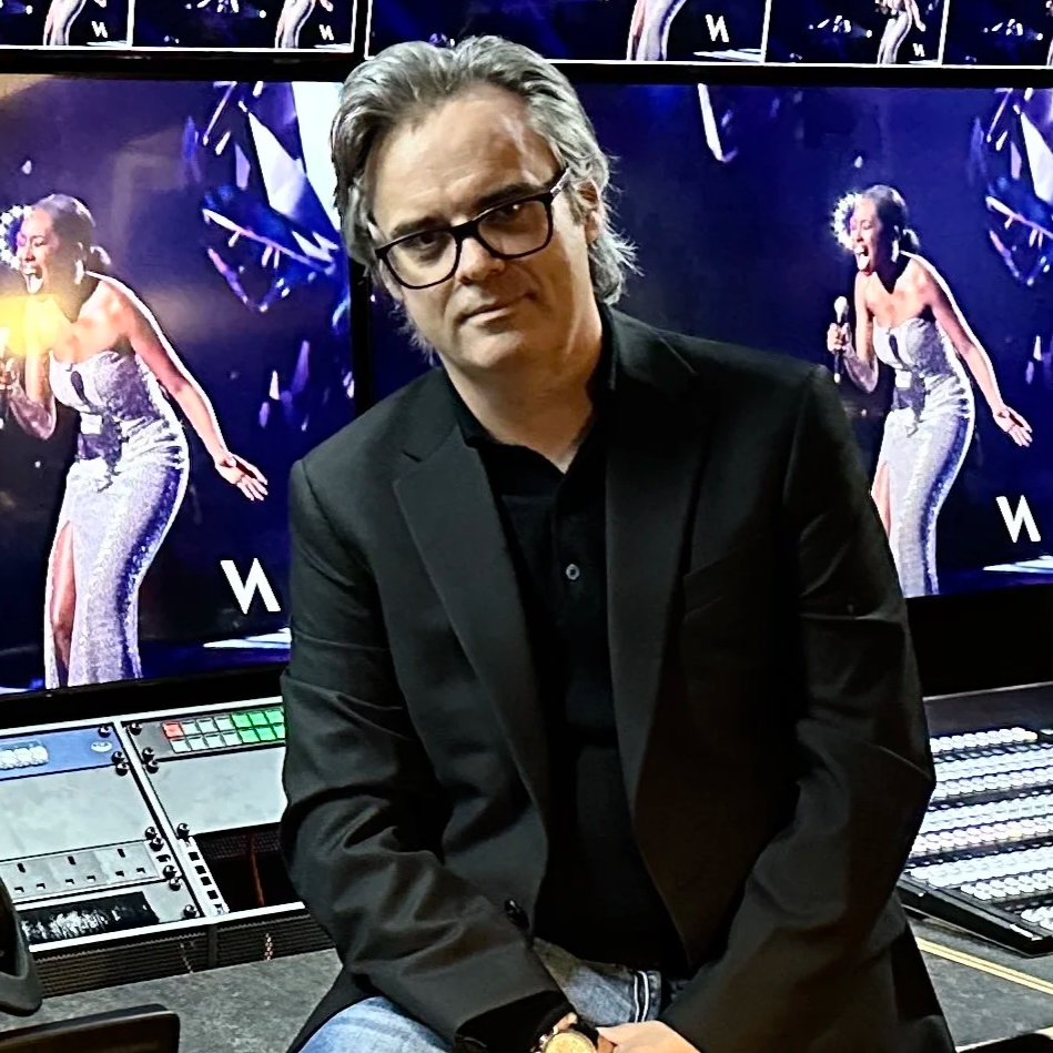 A man with gray hair and glasses wearing a black blazer sitting in front of multiple TV screens displaying a singer in a silver dress performing on stage.
