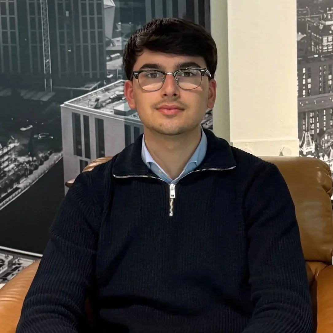 A young man with glasses, dark hair, and light skin, wearing a navy blue sweater with a zipper, sitting in a wooden chair in front of a cityscape black-and-white mural background.