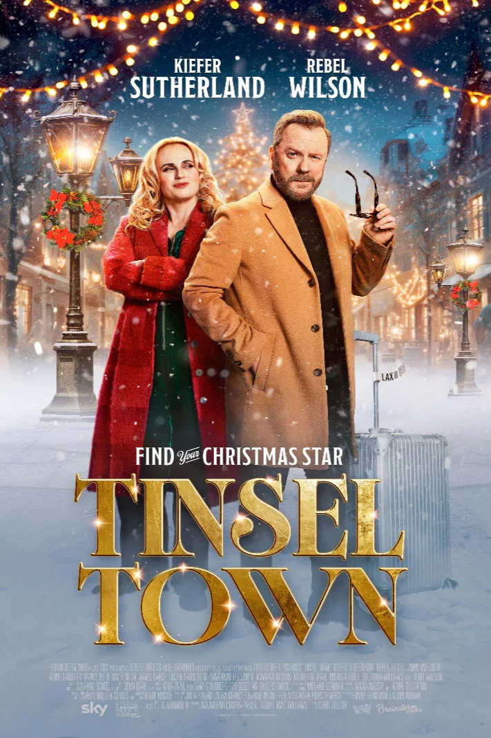 A festive holiday movie poster for 'Tinsel Town' featuring actors Kiefer Sutherland and Rebel Wilson standing on a snow-covered street decorated with Christmas lights and wreaths. Sutherland is holding glasses, Wilson is wearing a red coat, and there