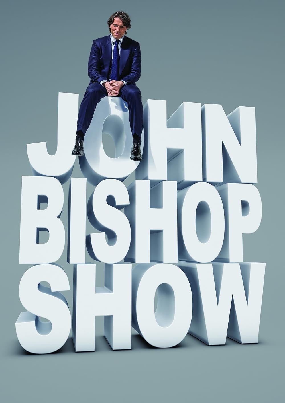 A man in a blue suit sitting on large white 3D letters spelling 'JOHN BISHOP SHOW' against a gray background.