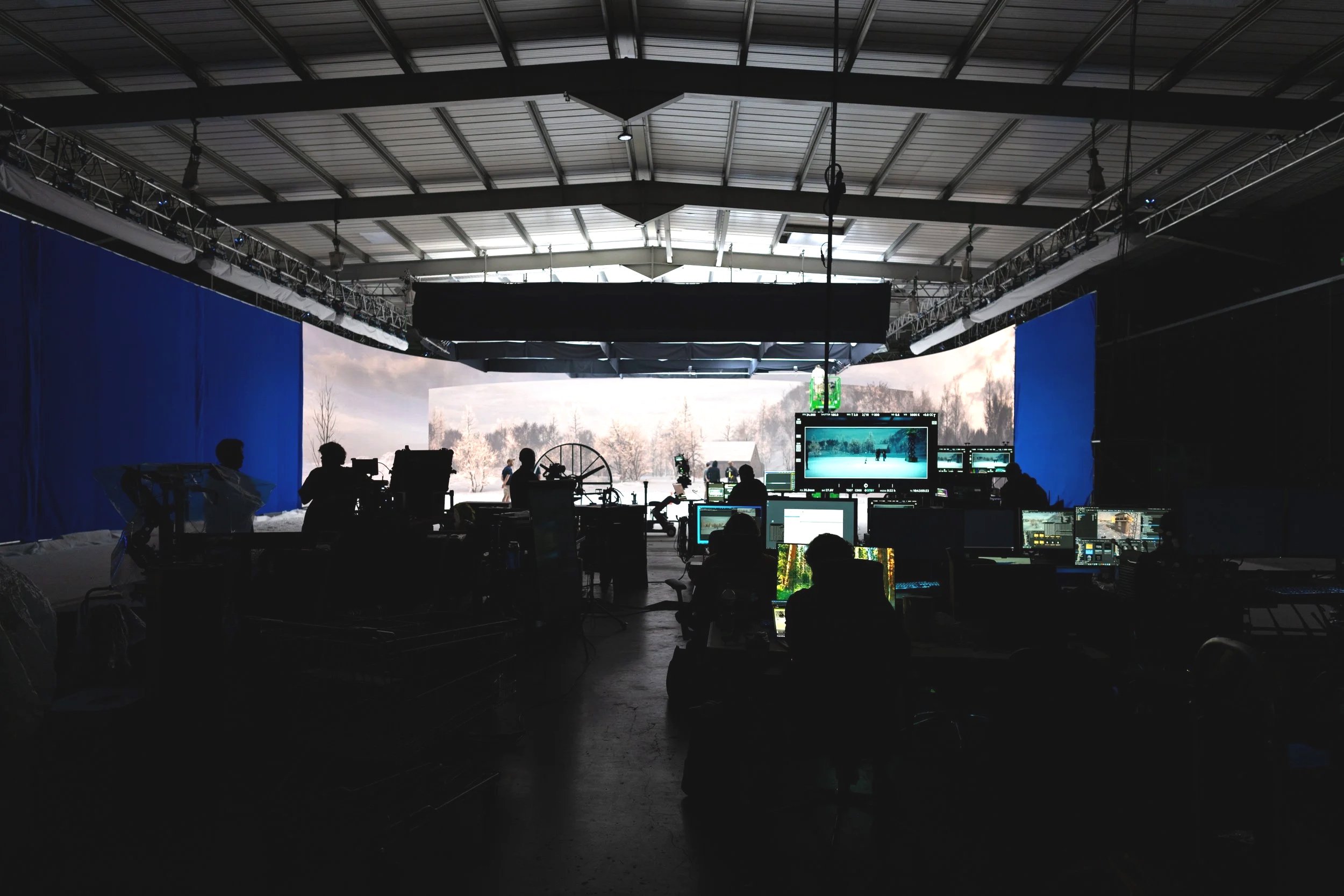 Film or photography studio with crew working on a shoot, multiple monitors displaying a winter landscape scene, film equipment, and crew silhouettes against the backdrop of an outdoor winter scene.