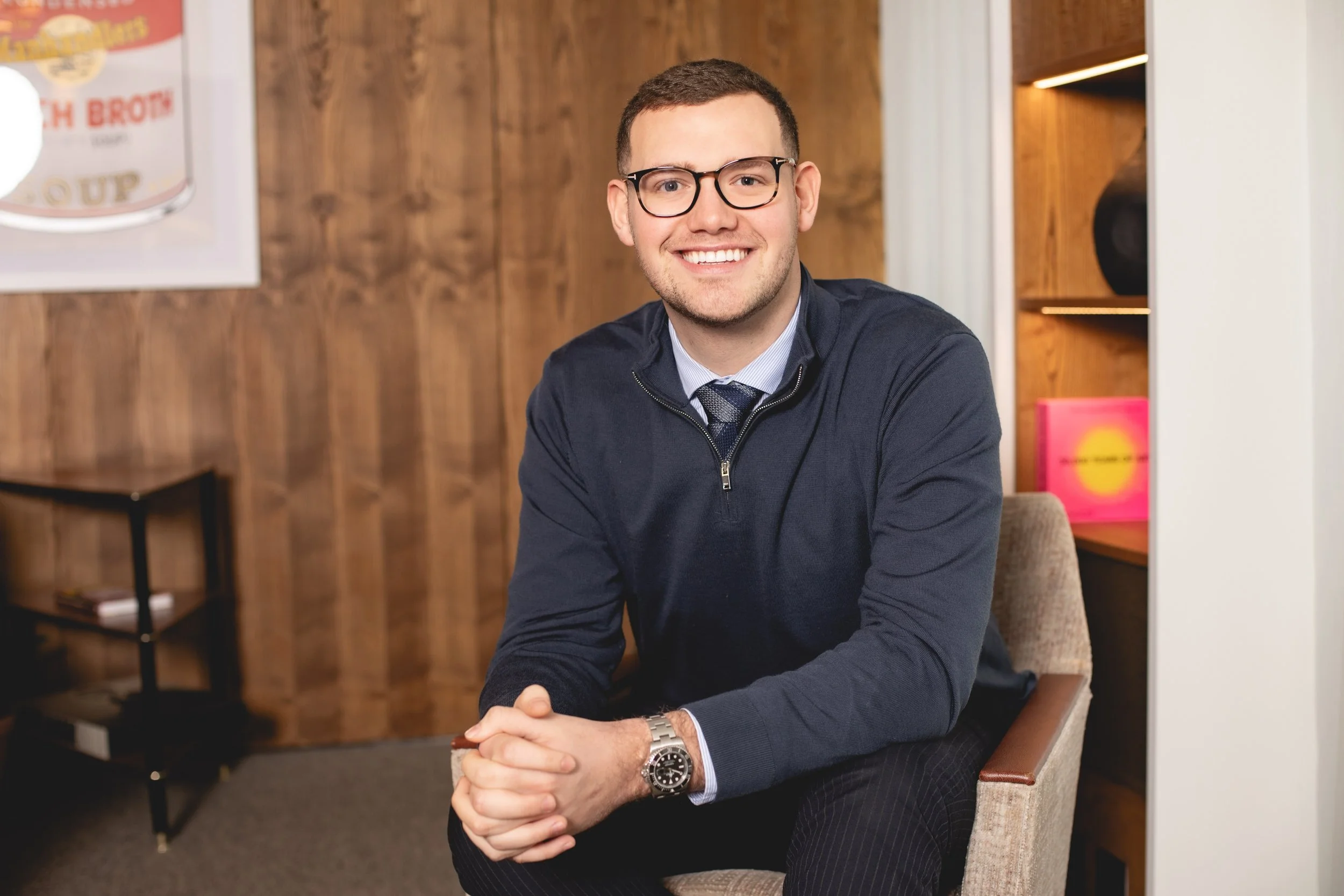 Smiling young man with glasses, wearing a dark sweater, tie, and watch, sitting in an office with wood-paneled walls and decorative shelves.