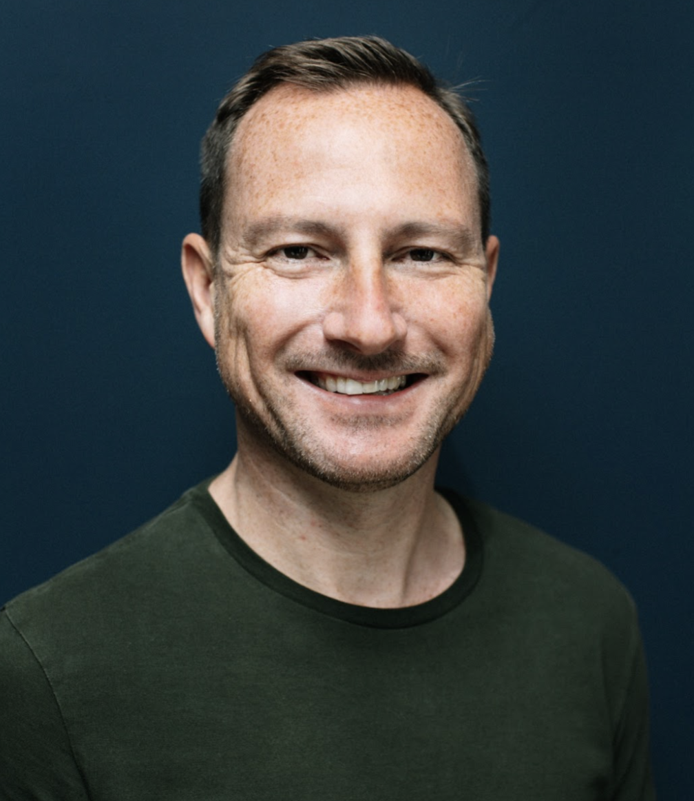 A smiling man with short brown hair, freckles, wearing a dark green shirt, standing against a dark blue background.