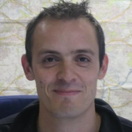 A young man with short, dark hair, slight facial hair, and a light smile, sitting in front of a map background.