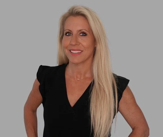 A woman with long blonde hair, wearing a black top, smiling against a gray background.