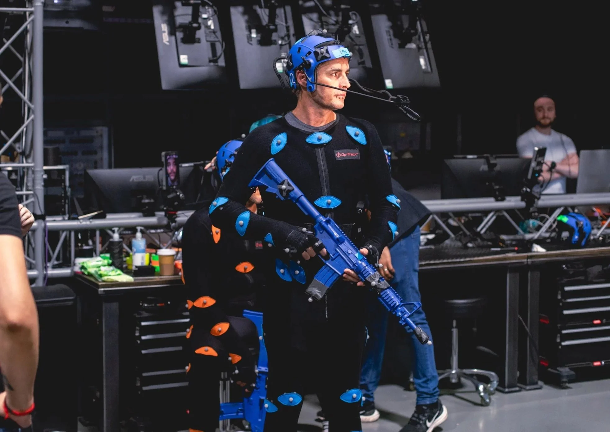 A man wearing motion capture suit with blue pads holding a blue rifle in a studio setting.