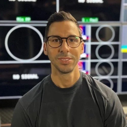 A man with dark hair, glasses, and a dark shirt, sitting or standing in front of a background with circular patterns and a screen displaying color bars.