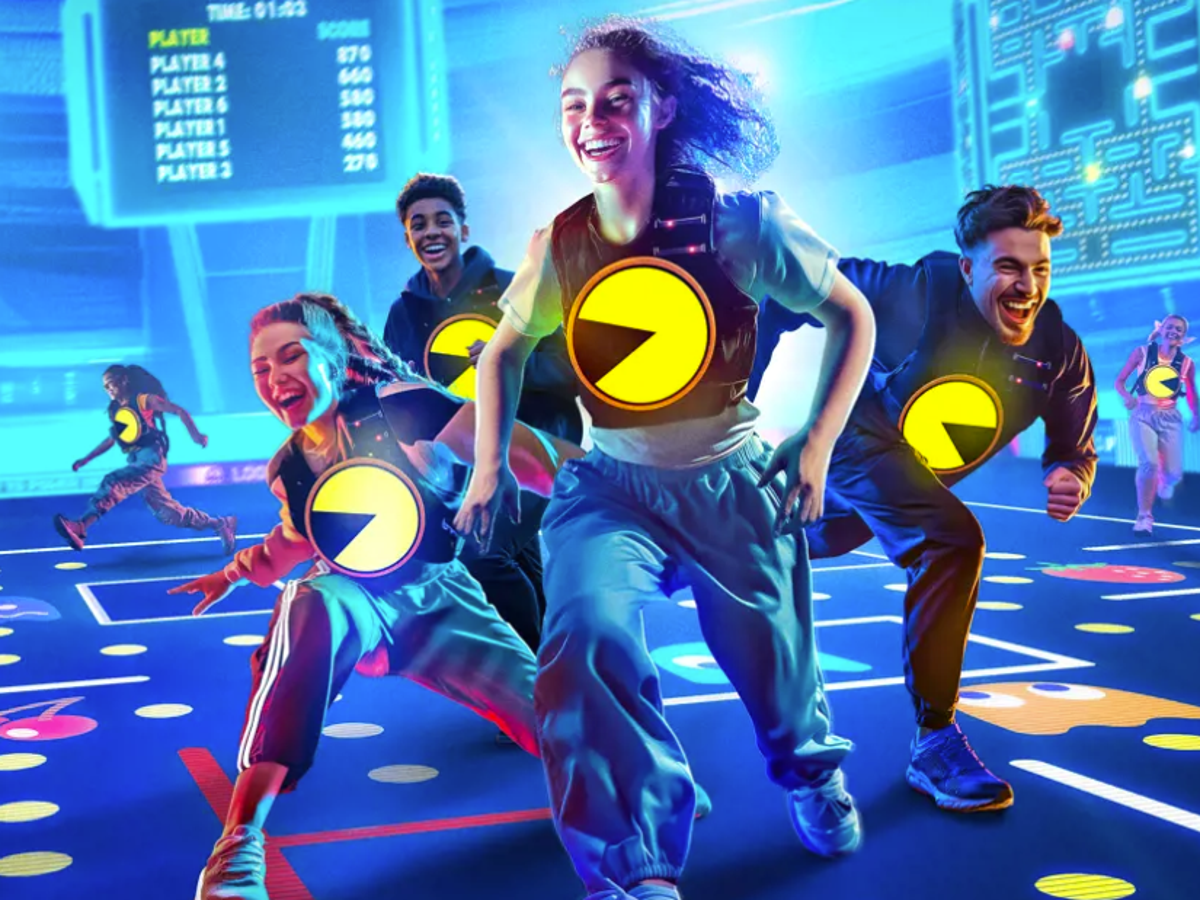 A group of five diverse young adults, wearing matching costumes with Pac-Man logo, dancing and playing in a vibrant, neon-lit arcade or gaming center. They appear to be having fun, with smiles and energetic movements.