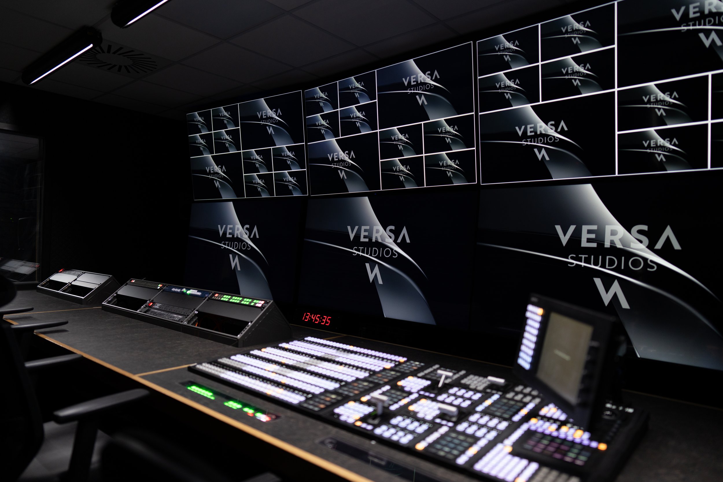 Dark control room with large digital screens displaying 'Verca Studios' logo, mixing consoles, and control panels illuminated with various lights, and a digital clock reading 13:45:35.