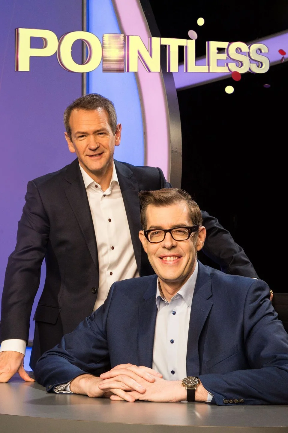 Two men in suits, one sitting at a desk with hands clasped and another standing behind him, smiling on a game show set named "Pointless" with colorful decorations and the show's logo in the background.