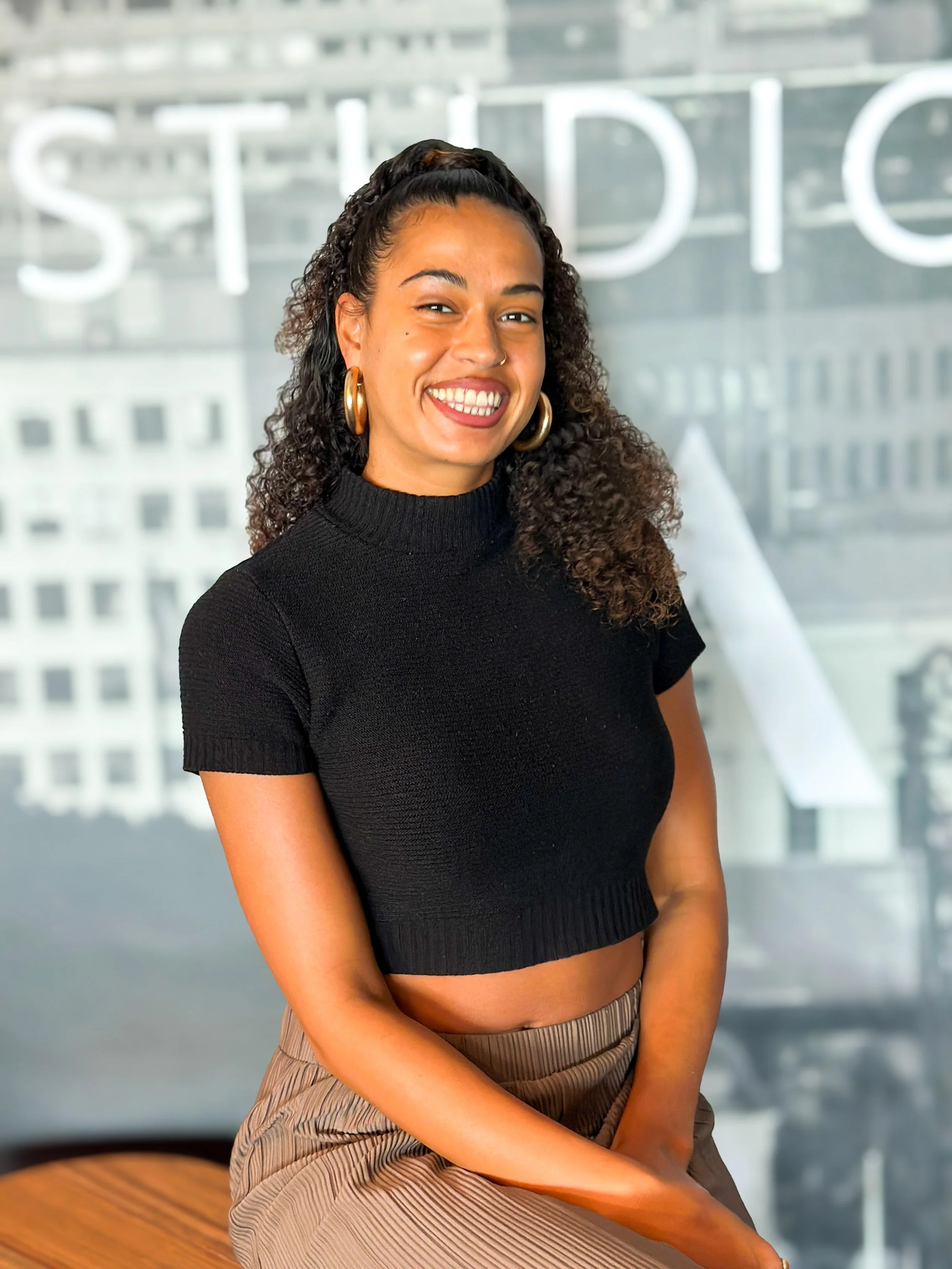 A smiling woman with curly hair, hoop earrings, wearing a black cropped sweater and beige pants, sitting in front of a window with an urban cityscape background.