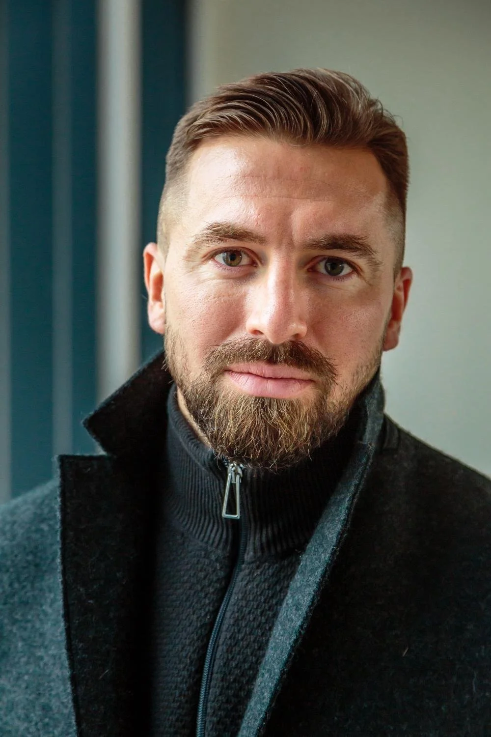 A close-up of a man with a beard and short brown hair, wearing a dark zip-up jacket with a high collar, standing indoors in front of a blurred background.