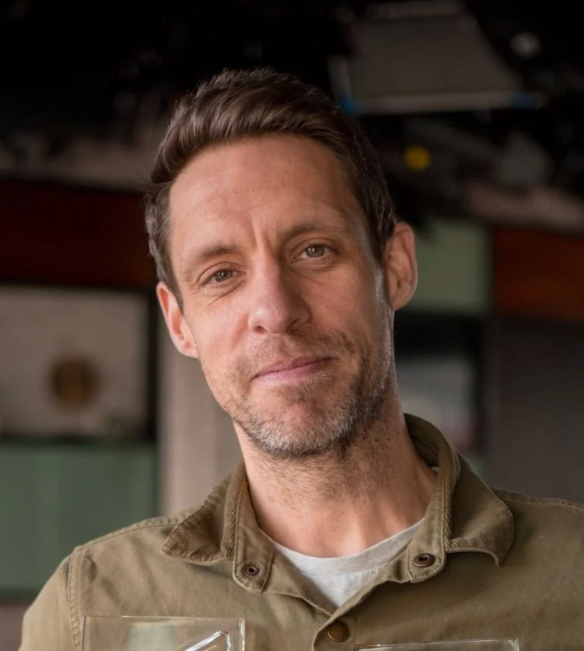 A man with short brown hair and light stubble on his face, wearing a khaki shirt, smiling softly inside a room with colorful storage boxes in the background.