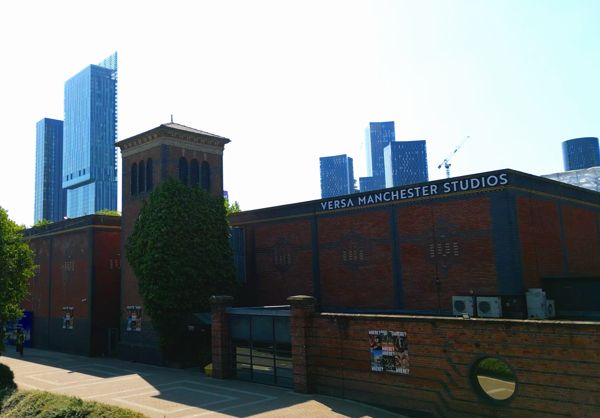 Photo of a brick building with the sign 'Versa Manchester Studios,' tall modern skyscrapers in the background, trees, and a parking lot in the foreground.