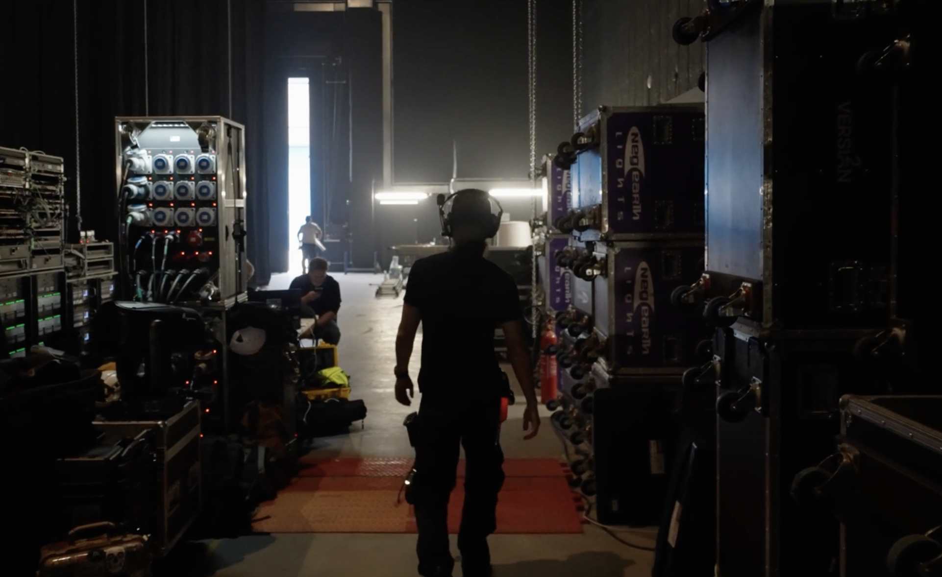 People working behind the scenes in a dark stage or production area, with equipment, power supplies, and cables visible, and natural light coming from open doors in the background.